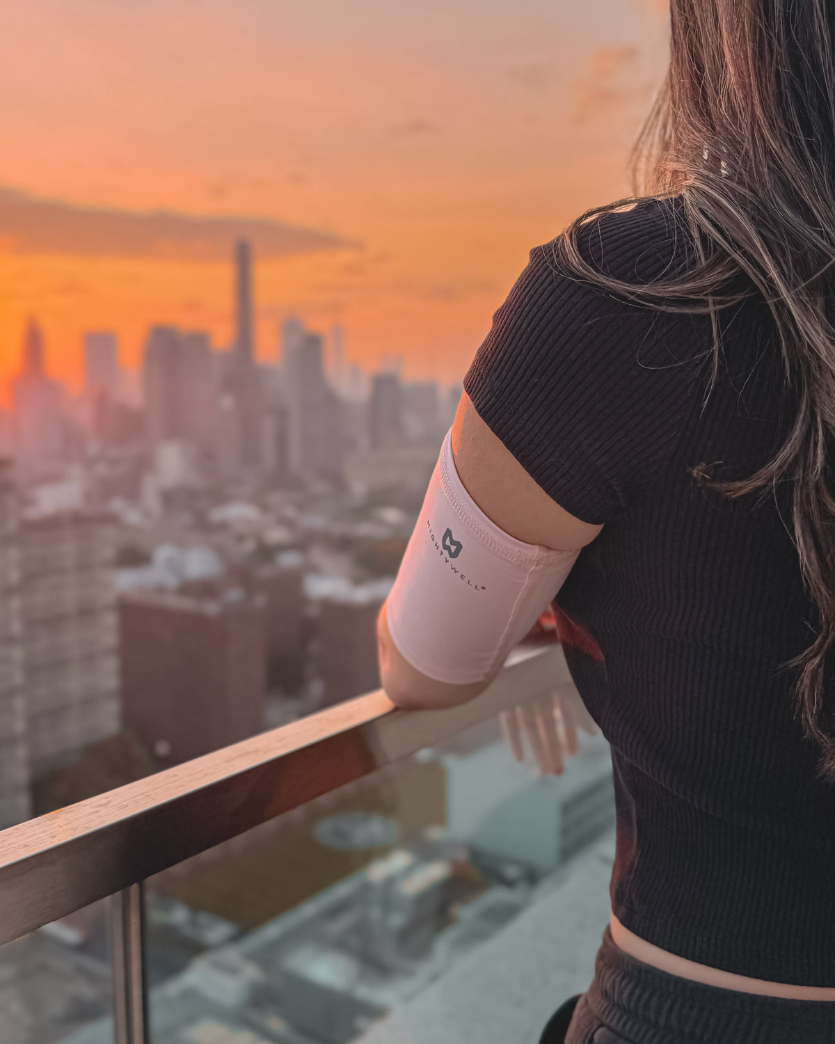 A woman in a black ribbed short-sleeve top stands on a balcony at sunset, wearing a light pink PICC line cover on her upper arm with a small logo.